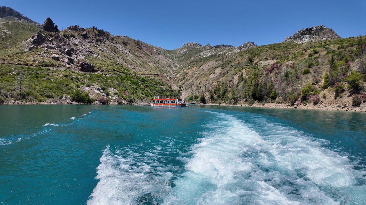 A wooden excursion boat moored below pine-covered canyon slopes on the turquoise water of Green Canyon, seen from the wake of a passing boat.